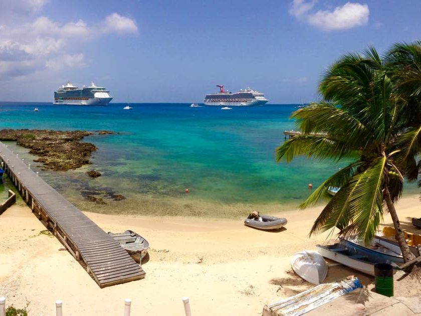 Cruise ships anchored off George Town, seen from a sandy shoreline with a wooden jetty and palms