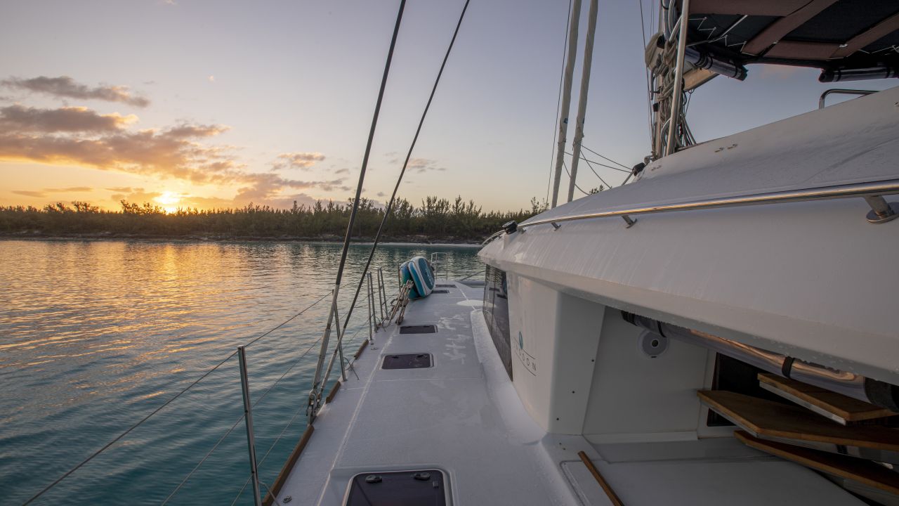 Side deck of a sailing catamaran at anchor as the sun sets behind a low, wooded shoreline