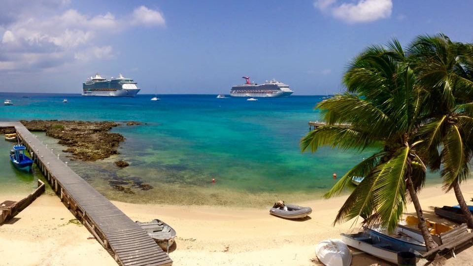 Two cruise ships anchored offshore beyond a wooden jetty, palms and a sandy shoreline in Grand Cayman